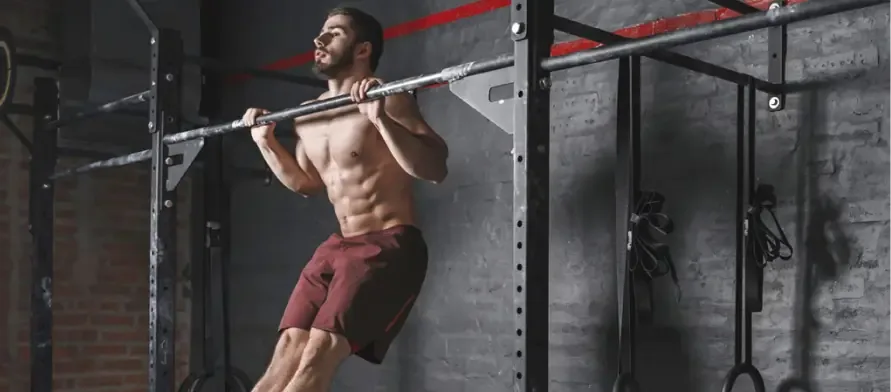 A shirtless man in red shorts doing a pull-up on a fixed rig bar in a dark commercial gym